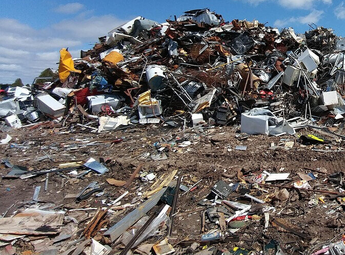 Worker in orange safety gear stands next to an orange dump truck dumping a huge pile of cardboard and paper waste inside a warehouse.