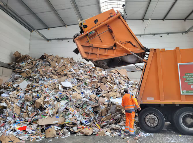 Worker in orange safety gear stands next to an orange dump truck dumping a huge pile of cardboard and paper waste inside a warehouse.